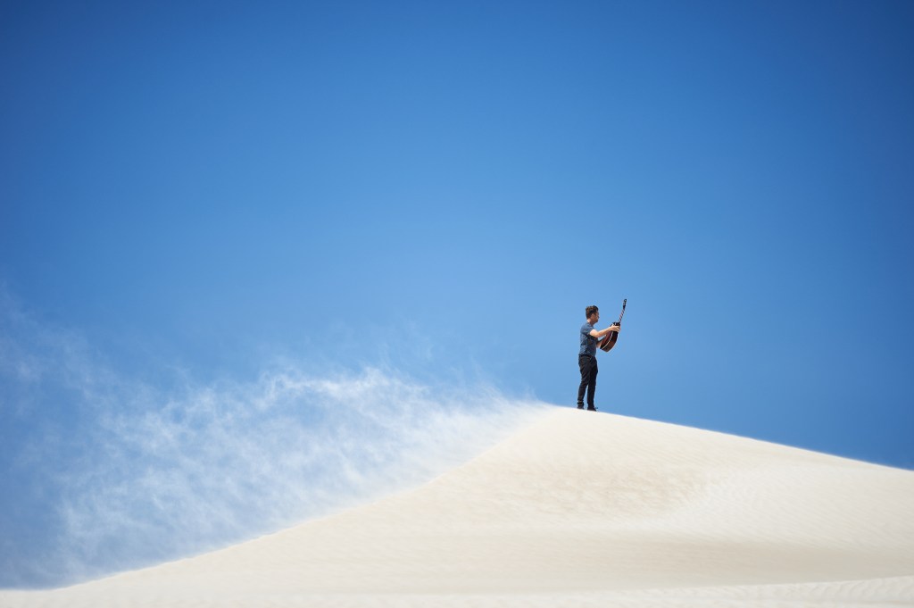 Stuart Orchard performing with an aeolian (wind-powered) guitar on a sand dune. A trail of sand blows behind him, and he is framed by a clear blue sky.